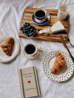 breakfast on a bed with coffee, croissants and blueberries next to a book