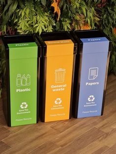 three different colored recycling bins sitting next to each other on a wooden floor with plants in the background