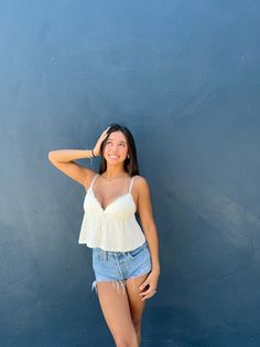 a beautiful young woman standing next to a blue wall wearing a white top and denim shorts