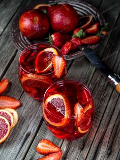 two glasses filled with red liquid and sliced strawberries next to a knife on a wooden table