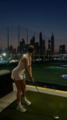 a woman is playing golf in front of a city skyline at night with the lights on