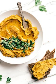 a white bowl filled with hummus next to two slices of bread and green leaves