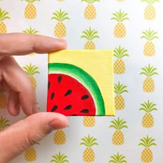 a hand holding a piece of paper with a watermelon painted on it and pineapples in the background