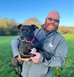 a man holding a black dog in his arms on a green field with trees and blue sky