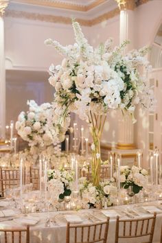 A large, round table adorned with a white tablecloth and a stunning floral centerpiece featuring cascading white orchids, hydrangeas, and delphinium. The centerpiece is surrounded by candles and crystal glassware. Wedding White Flowers, Floral Centerpieces Wedding, Glamorous Room, Plaza Wedding, Grand Ballroom, Centerpieces Wedding, Breathtaking Wedding, Wedding Decor Inspiration