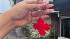 a hand holding a red and green crocheted flower keychain in front of a house