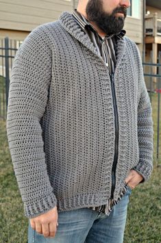 a man with a beard wearing a gray sweater and jeans stands in front of a house