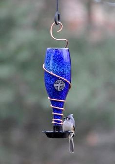 a blue glass bird feeder hanging from a metal hook with a small bird on it