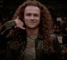 a man with long curly hair wearing a leather jacket and pointing to his left side