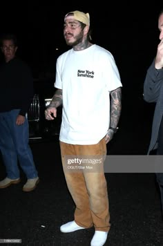 tattooed man in white t - shirt and tan pants standing outside at night with other people