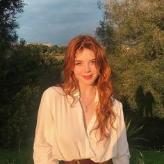 a woman with long red hair wearing a white shirt and brown belt standing in front of trees