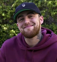 a man with a beard wearing a purple hoodie and a black hat smiles at the camera