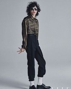 a man with curly hair and sunglasses standing in front of a grey background wearing black pants