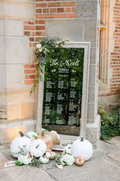 a wedding seating chart with white pumpkins and greenery on the ground in front of a brick building