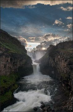 an image of a waterfall in the middle of some rocks and grass with clouds above it