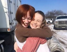 two women hugging each other in front of a truck and snow covered road with trucks behind them