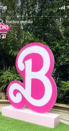 a pink and white b sign sitting on top of a lush green field with trees in the background