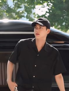 a young man standing in front of a car wearing a black shirt and baseball cap