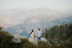 a man and woman holding hands on top of a mountain with mountains in the background