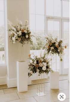 three white vases with flowers and greenery on them in front of large windows