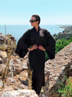 This gothic long kimono robe is a striking piece that blends dark elegance with luxurious comfort. Made from black satin, this kimono robe for women exudes a sense of sophistication and mystique, perfect for those who appreciate a gothic aesthetic. The bell-sleeve dress design adds a touch of drama and flair, enhancing the overall allure of the robe. Ideal as a gift for your wife, this elegant dressing gown is both practical and stylish, making it a cherished addition to her wardrobe.  ◌ SIZE: - Evening Kimono With Kimono Sleeves, Black Open Front Evening Kimono, Black Long Evening Robe, Long Black Evening Robe, Black Samurai Kimono With Kimono Sleeves, Long Black Summer Abaya, Elegant Black Summer Abaya, Black Maxi Length Kimono, Elegant Long Black Robe