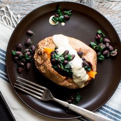 a black plate topped with a baked potato covered in beans and greens next to a fork