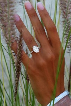 a woman's hand with a diamond ring on her finger in front of some tall grass