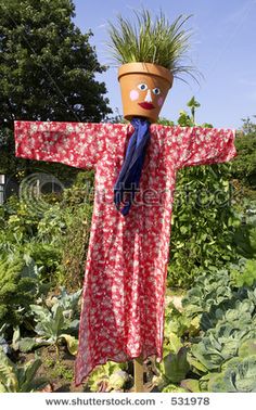 a scarecrow in a red dress with a flower pot on his head standing in a garden