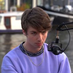 a young man sitting next to a microphone in front of a boat on the water