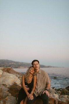 a man and woman are sitting on rocks by the water with their arms around each other