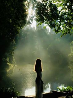 a woman standing in front of a body of water with the sun shining through trees