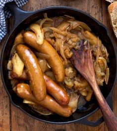 a pan filled with sausages and onions on top of a wooden table next to bread