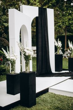 two black vases with white flowers are sitting on the grass in front of an arch