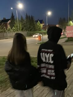 two people are walking down the sidewalk at night, one is wearing a black hoodie