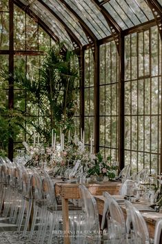 a long table with clear chairs and lots of greenery in the background is surrounded by tall glass windows