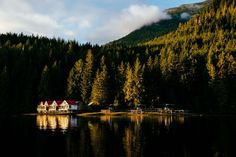 a house on the shore of a lake surrounded by pine trees and mountains in the background