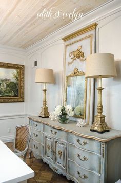 an antique dresser with two lamps and paintings on the wall in a white dining room