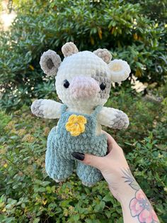 a hand holding a small stuffed animal in front of some bushes and trees with flowers on it