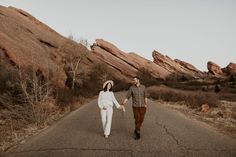 a man and woman holding hands walking down an empty road in front of large rocks