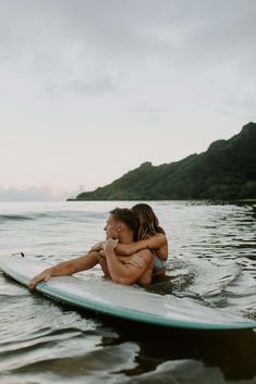 two people are sitting on a surfboard in the water, one is hugging the other