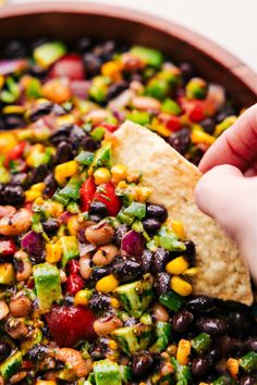 a hand dipping a tortilla chip into a bowl filled with black beans, corn and avocado