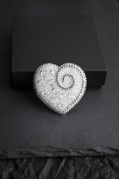 Beautiful and tender embroidered brooch in the shape of a white and silver heart. It is embroidered with seed beads, Czech crystal faceted beads and rhinestone chain. The reverse side is covered with silver faux leather. Each element of brooch is securely sewn by hand. Japanese smooth pin for a secure fit without damaging clothes.  It will decorate your look or can become an unforgettable gift for a dear person. SIZE: Length - 1.9 inches / 4.8 cm Width - 2 inches / 5.2 cm Weigh - 7 gr Will come Elegant Silver Heart Brooches, Elegant Silver Heart-shaped Brooches, Elegant Silver Beaded Brooches, Elegant Silver Heart Brooch, Elegant Silver Heart-shaped Brooch, Elegant Heart-shaped Wedding Brooch, Silver Embellished Wedding Brooches, Embellished Round Beads Jewelry Gift, Gift Jewelry With Embellished Round Beads