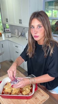 a woman is cutting up food in a red dish on a wooden table with white cabinets