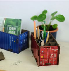 a container with pens and pencils in it on a table next to a potted plant