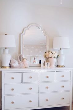 a white dresser topped with lots of drawers next to a wall mounted mirror and two lamps