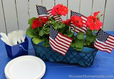 red flowers in a blue basket with american flags and straws on the table next to a white plate