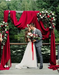 a bride and groom kissing in front of an outdoor ceremony arch with red drapes