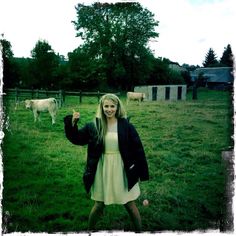 a woman in a white dress and black jacket posing for the camera with cows behind her