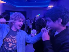 two young men standing next to each other holding chopsticks in their mouths and laughing