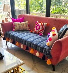 a pink couch with colorful pillows on it in front of a large window and table